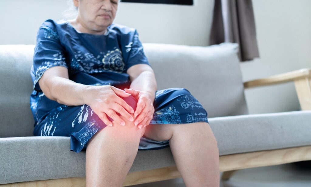 une personne âgée assise dans son canapé qui a des douleurs articulaires au genou